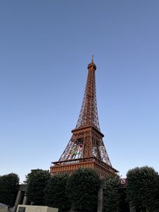 The Eiffel Tower with Olympic rings attached, set against a clear blue sky, with green trees at its base and a partially visible no-parking sign below.
