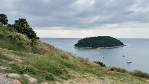 A view from a grassy hillside overlooking a calm sea. In the distance, there is a small, lush island surrounded by water, with several white sailboats anchored nearby