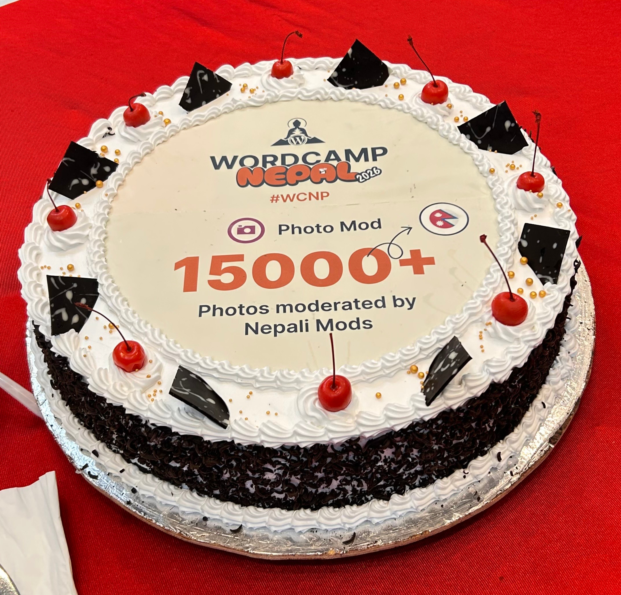 A round cake with white frosting and a WordCamp Nepal 2026 label, decorated with chocolate shards, red cherries, and golden sprinkles, placed on a red tablecloth.