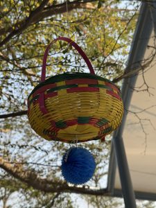 A colorful woven basket hanging from a branch, with a pink handle and a decorative blue pom-pom below it.
