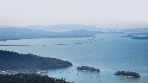 Serene coastal landscape featuring calm waters reflecting a soft blue hue, surrounded by lush green hills and small islands. Between the land and the horizon, several sailboats are gently anchored, creating a peaceful scene. In the distance, a series of peninsulas stretch across the water, leading toward hazy mountains that fade into the sky.