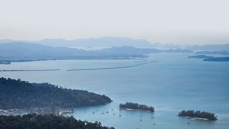 Serene coastal landscape featuring calm waters reflecting a soft blue hue, surrounded by lush green hills and small islands. Between the land and the horizon, several sailboats are gently anchored, creating a peaceful scene. In the distance, a series of peninsulas stretch across the water, leading toward hazy mountains that fade into the sky.