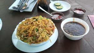 A white bowl of stir-fried noodles with chicken and vegetables sits on a saucer. Next to it is a bowl of dark broth with herbs, small containers of chili paste and sauce, and lime slices on a plate in the background.