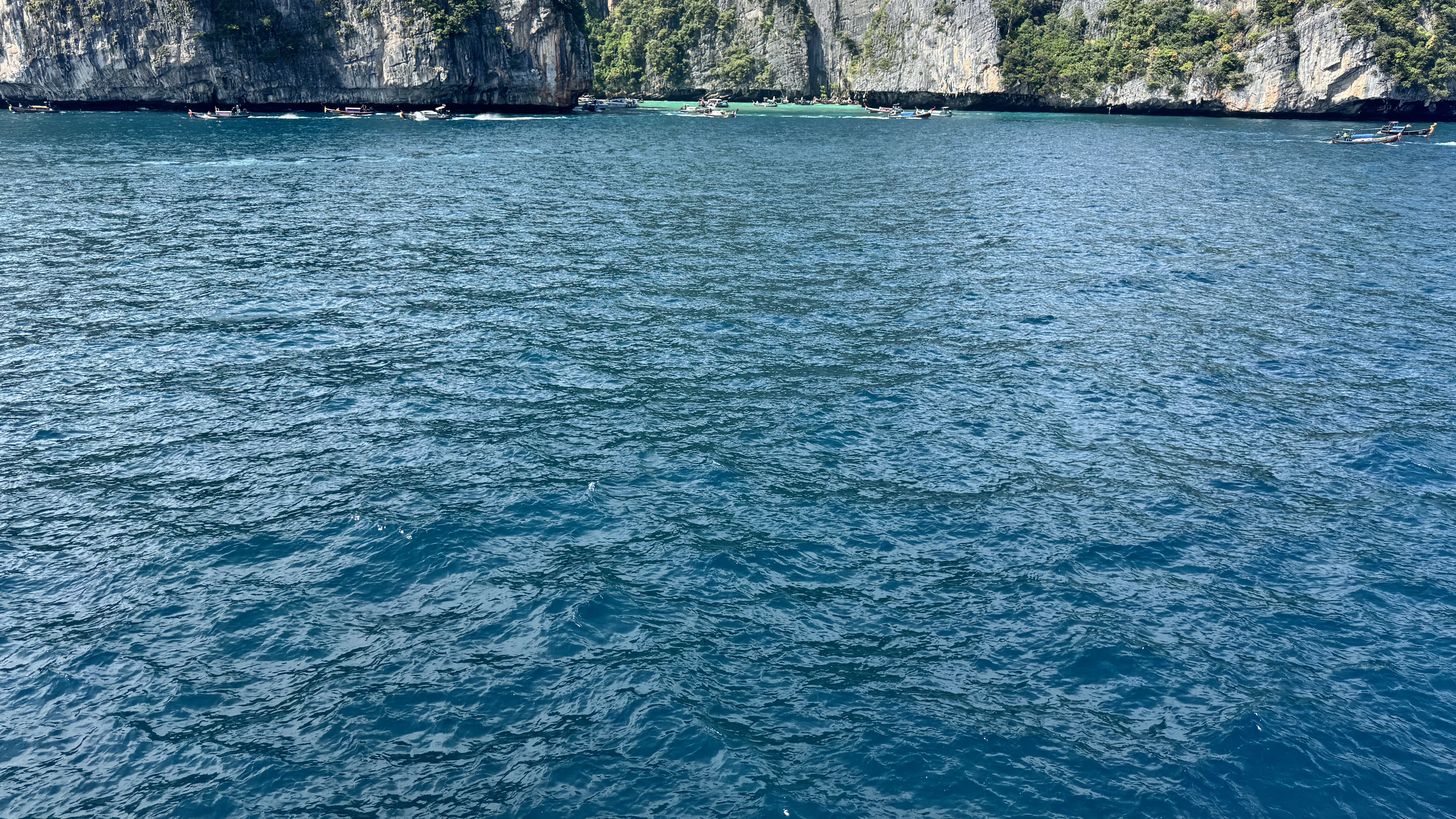 A view focusing on the rippling surface of deep blue and turquoise ocean water, with a distant glimpse of limestone cliffs and boats anchored along the top edge.