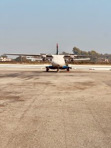 A symmetrical, head-on view of a white twin-turboprop aircraft parked on an airport runway. The nose is protected by a white weather cover, and the propellers are visible against a clear sky with a distant treeline and small buildings.