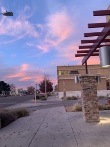 A sidewalk scene at sunset featuring a movie theater with the name “Bravo!” glowing in yellow lights, set against a pink and blue sky with wispy clouds, with nearby trees, parked cars, a stone pillar, and modern light fixtures adding to the urban atmosphere.