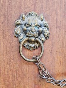 A detailed lion-shaped metal door knocker with a chain, fixed on a wooden door. The texture and craftsmanship stand out clearly in Kozhikode, Kerala.