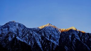 A simple Manali hill view with snow-covered mountains under a clear blue sky, as sunlight softly lights up one peak.