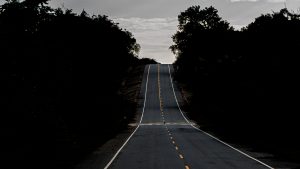 A centered, symmetrical view of a long gray asphalt road with a dashed yellow line, stretching uphill into the distance, flanked by dense dark forest silhouettes under a cloudy, overcast sky.