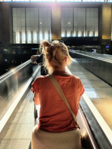 A woman with light, tousled hair is seated on a moving walkway, facing away from the camera. She wears a casual orange top and has a light-colored bag beside her.