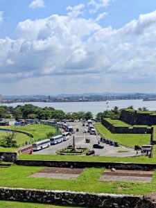 View from the ramparts of Galle Fort in Sri Lanka showing a busy roundabout with buses and tuk-tuks, green fort walls, and the sea with a distant shoreline under a partly cloudy sky.
