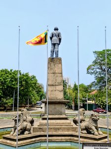 Rear view of the statue of D. S. Senanayake at Independence Memorial Hall in Colombo, Sri Lanka, standing on a tall stone pedestal, surrounded by lion sculptures and a circular fountain, with the Sri Lankan flag flying beside it and city surroundings in the background.
