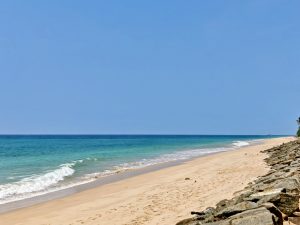 Sandy beach in Bentota, Sri Lanka, with gentle waves of the Indian Ocean meeting the shore, clear blue sky above, and a rocky embankment running along one side.
