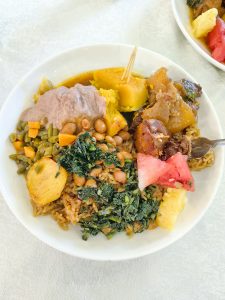A white bowl filled with a variety of colorful foods. The dish includes yellow plantains, a smooth grayish-purple sauce, a mix of steamed greens with chopped vegetables, rice with beans, and several pieces of meat