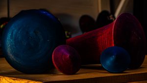 Four geometric objects with teal and magenta metallic finishes sit on a wooden table in a dark setting.