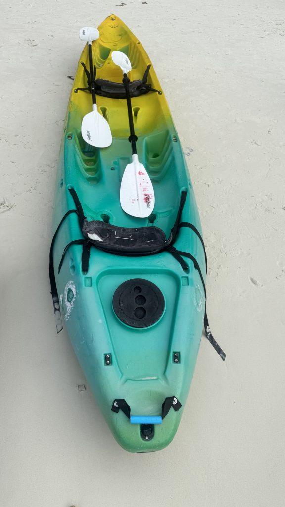 A top-down view of a yellow and green sit-on-top kayak with two white paddles resting on a white sand beach.