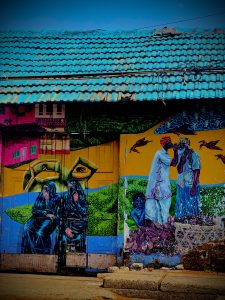 Painting on the streets of Gujarathi street Calicut.  The painting is a brightly colored street scene on the side of a building.  The roof color nearly matches the sky. 