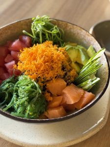 A colorful poke bowl with salmon, tuna, seaweed, avocado, cucumber, and carrots in a ceramic bowl on a wooden table.