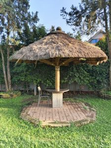 A rustic outdoor gazebo with a thatched roof and wooden pillar, covering a small round table and chair on a circular patio, surrounded by green grass, shrubs, and tall trees in a peaceful garden