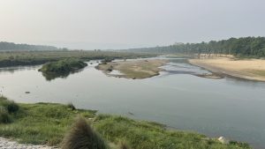 

A serene landscape featuring a winding Mahakali river with calm water reflecting the sky. Lush green grass lines the riverbanks, and small islands of grass and sand emerge from the water.