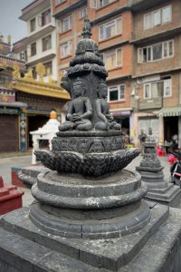 A close-up view of a dark stone statue featuring two seated figures, each depicted in a meditative pose. The figures are flanked by intricate carvings on the statue's pedestal, which rests on a lotus-like base.