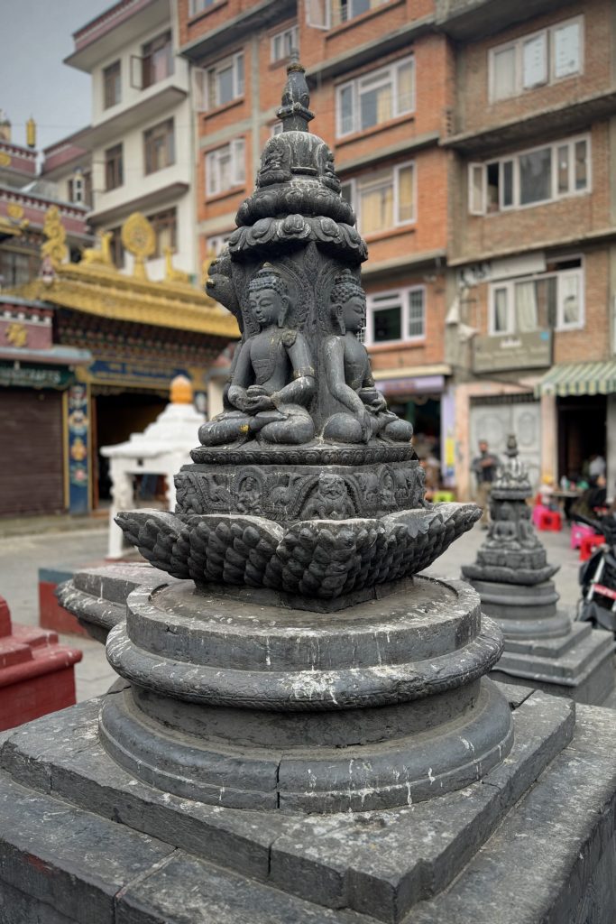 A close-up view of a dark stone statue featuring two seated figures, each depicted in a meditative pose. The figures are flanked by intricate carvings on the statue’s pedestal, which rests on a lotus-like base.