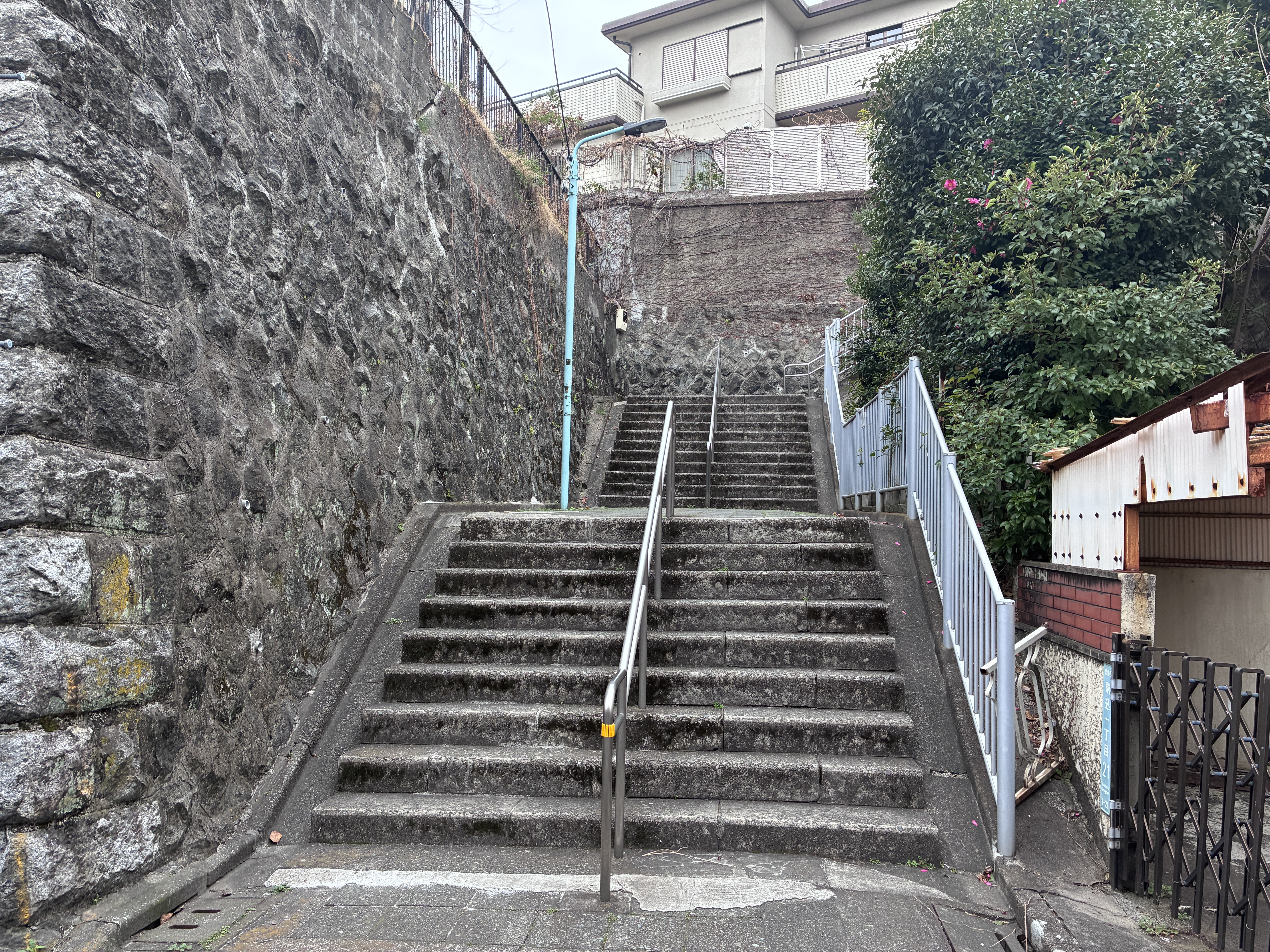 A set of concrete stairs leads up between two stone walls. The left wall is made of rough, dark stones, while the right side features a white railing and lush greenery.