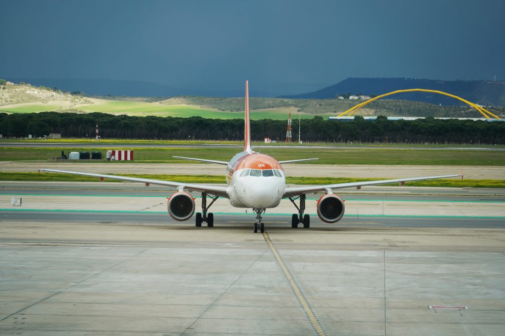 A commercial airplane is positioned and facing the viewer, showcasing a white body with orange accents. The aircraft is situated on a runway with visible markings and green taxiways.