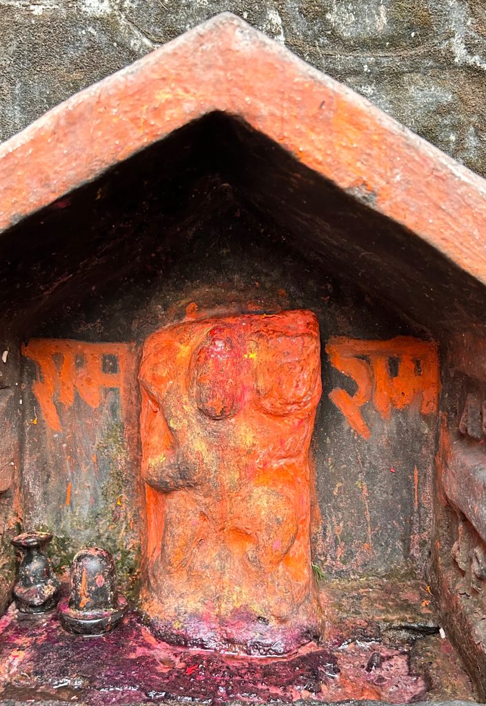 A small shrine is carved into a stone wall, featuring a vivid orange idol with smooth curves and a slightly abstract form, possibly representing a deity.