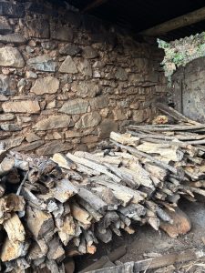 
A rustic stone wall is partially visible in the background, made of various sized and shaped stones with a natural earthy color. In the foreground, there is a neatly arranged pile of firewood consisting of split logs and branches, showcasing a mixture of textures and colors typical of dried wood