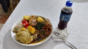 A plate of mixed food is displayed, featuring a variety of items including mashed starchy vegetables, pieces of meat, leafy greens, a portion of rice, and chopped fresh tomatoes. The plate is accompanied by a bottle of soda and a fork, all set on a white tablecloth.