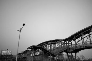 A gracefully curved wooden pedestrian bridge arches against a clear sky in this black and white urban scene. Below the elevated structure, a street lamp and the edge of a nearby building provide context to the bustling city environment.