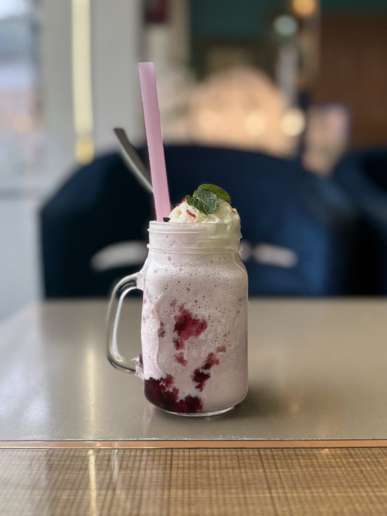 Strawberry milkshake in a mason jar topped with whipped cream, mint leaves, and a pink straw on a café table.