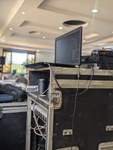 A close-up view of a laptop placed on a dark, textured audio equipment rack. The laptop has multiple cables connected to it.
