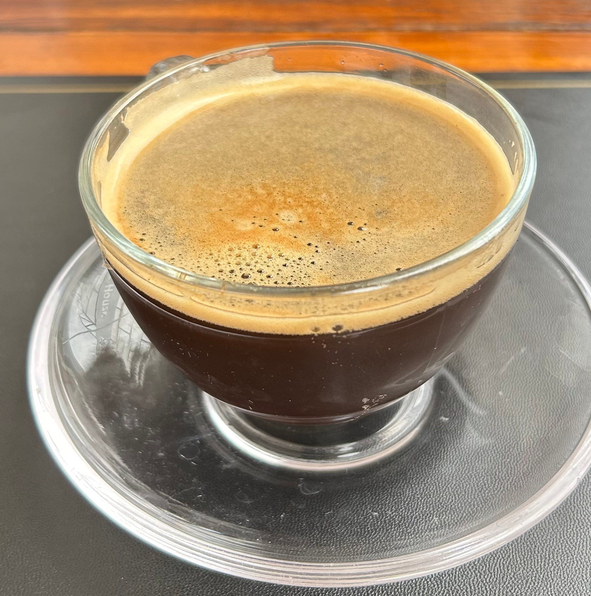 A glass cup of dark coffee sits on a small glass saucer.