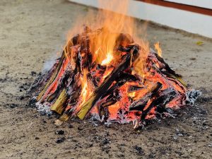 A large bonfire burning brightly with flames reaching upward, surrounded by charred logs and embers on a sandy surface. 