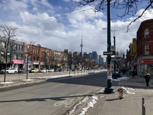 At the corner of Oxford St. and Spadina Ave. (Kensington-Chinatown, Toronto) in late winter, the CN tower is visible above the bare trees in the boulevard median. Long shadows fall over the streets and bike lane where a little snow remains below the historic three-storey brick buildings that line Spadina.
