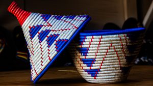 A traditional handwoven basket with a matching conical lid, featuring blue, red, and cream geometric patterns, on a wooden surface.