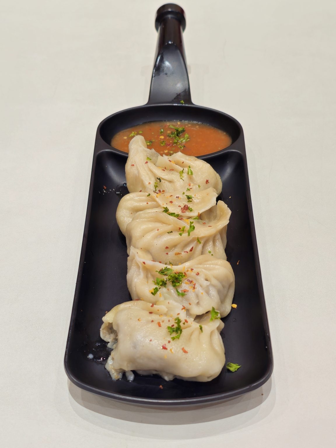 Steamed dumplings (momos) served in a black dish with spicy dipping sauce. The soft texture and simple presentation make the dish look delicious in this photo taken in Mavoor Road, Kozhikode.