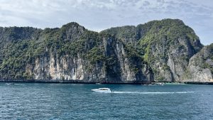 A view from a moving boat showing steep limestone cliffs covered in dense green vegetation rising from the blue ocean, with a white speedboat passing by.