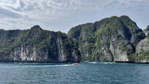 A scenic seascape featuring towering, forested limestone cliffs with jagged rock formations, surrounded by numerous tour boats in the bright blue water.