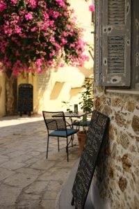 This vertical photograph captures a charming outdoor café scene with a black chalkboard menu leaning against a rustic stone wall. In the background, a small table and chair sit in the warm sunlight, framed beautifully by a large, vibrant pink bougainvillea plant in full bloom.
