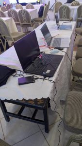 A table covered with a white lace cloth displays three open laptops, not powered on, one is alongside a portable battery pack and a computer mouse. Various cables are connected to the devices. Behind the table, several empty chairs are arranged, and a stage with lighting can be seen in the background. 
