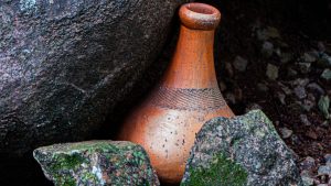 A long-necked terracotta clay pot with etched line patterns nestled between large, textured grey boulders and smaller moss-covered rocks on a dark soil ground.
