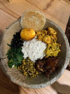 A traditional Nepali food platter served in a leaf bowl, featuring beaten rice in the center surrounded by various items including a fried egg, spicy potatoes, greens, beans, and roasted meat.