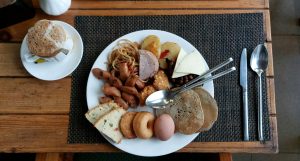 A round white plate is filled with a variety of breakfast foods, including spaghetti, sliced sausages, a slice of meat, fried potato slices, pieces of cheese, black beans, bread, a donut, a soft-boiled egg, and a stack of pancakes.