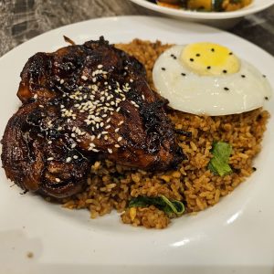 A plate of food featuring grilled chicken glazed with a dark sauce, sprinkled with sesame seeds, resting on a bed of seasoned rice.
