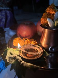 A decorative tray with red apples, marigold flowers, and a small brass pot, alongside a glowing oil lamp on a leaf, set in a soft nighttime light.