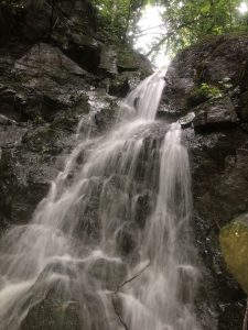 A cascade of water flows over dark, moss-covered rocks, framed by lush greenery above. The soft, white water contrasts with the textured stone, creating a serene and natural scene. Santa Cruz, Guanacaste
