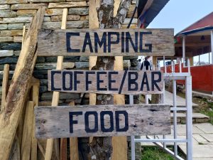 Rustic wooden signpost attached to a tree pointing to “Camping,” “Coffee/Bar,” and “Food,” with a stone building and walkway behind it.